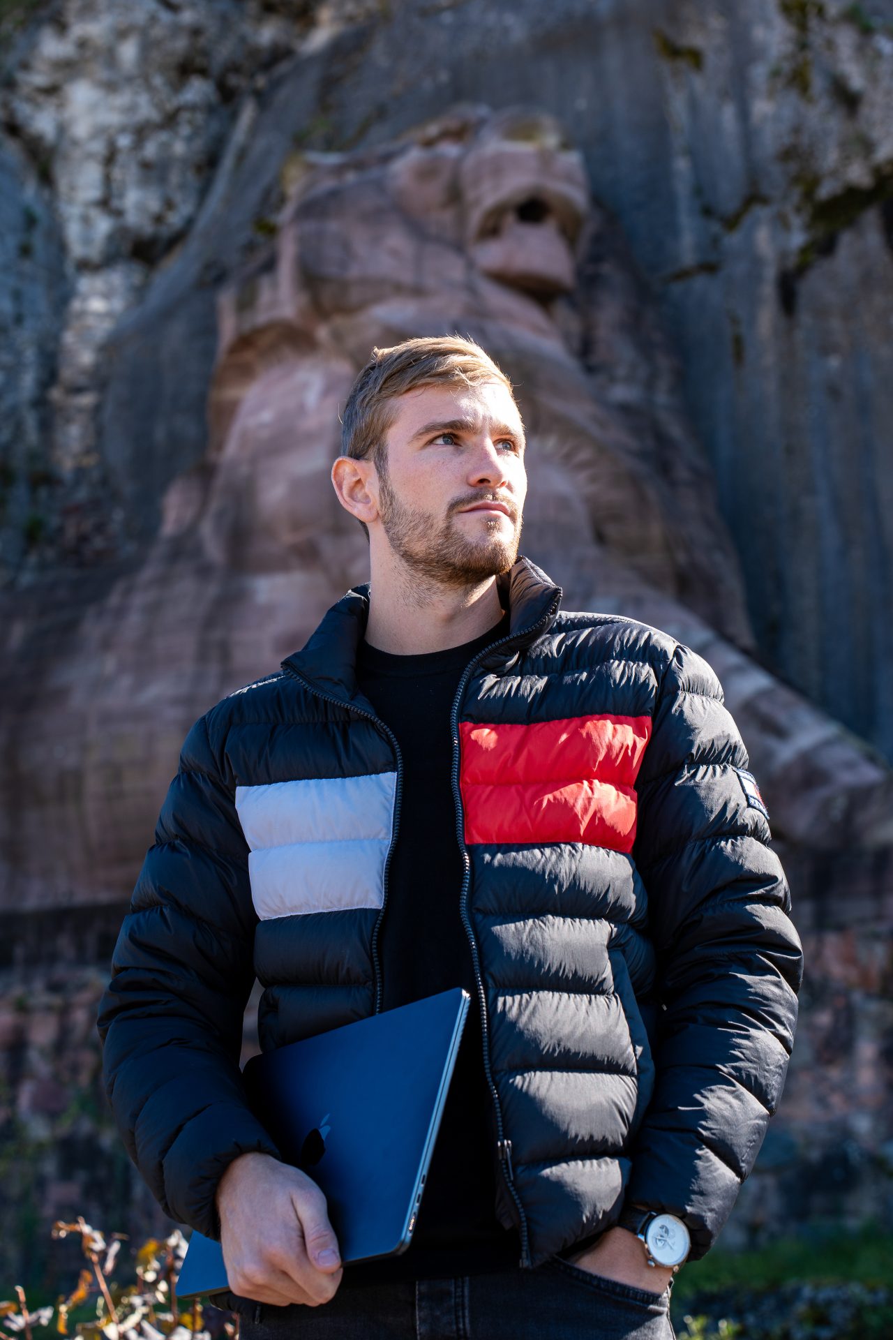 Alban portant un blouson se tient à l'extérieur, près d'une rambarde métallique, et utilise un ordinateur portable. Des falaises rocheuses et du feuillage sont visibles à l'arrière-plan sous un soleil éclatant.