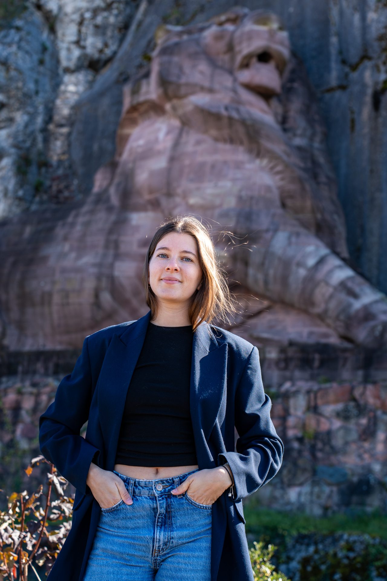 Margaux portant un blouson se tient à l'extérieur, près d'une rambarde métallique, et utilise un ordinateur portable. Des falaises rocheuses et du feuillage sont visibles à l'arrière-plan sous un soleil éclatant.