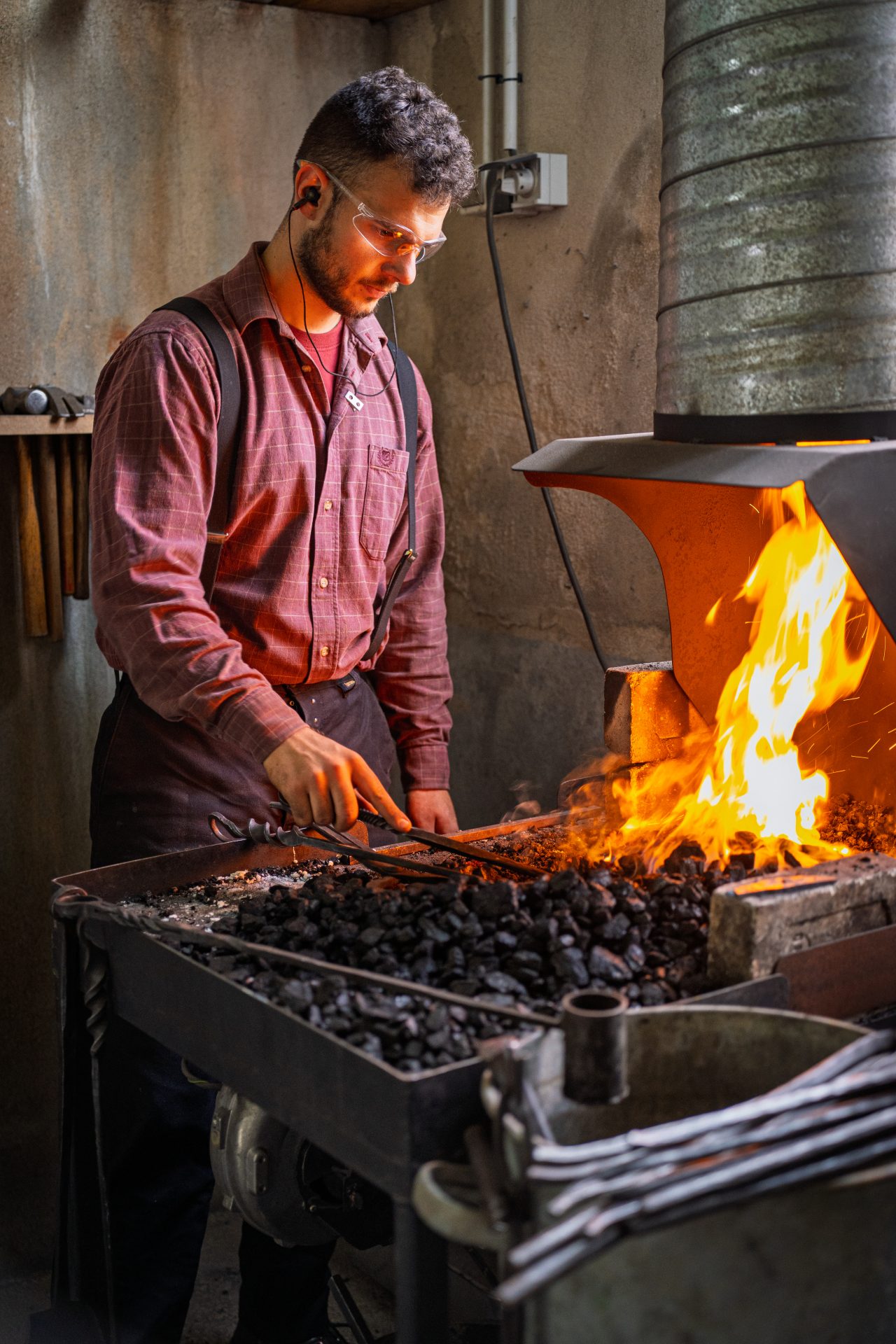 Un forgeron portant des lunettes de sécurité façonne le métal avec des pinces au-dessus d'une forge brillante et chaude dans un atelier, entouré d'outils et de charbon brûlant.